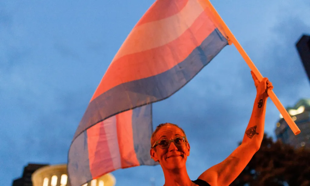 Una persona sostiene una bandera trans, ambas bañadas en un brillo naranja.
