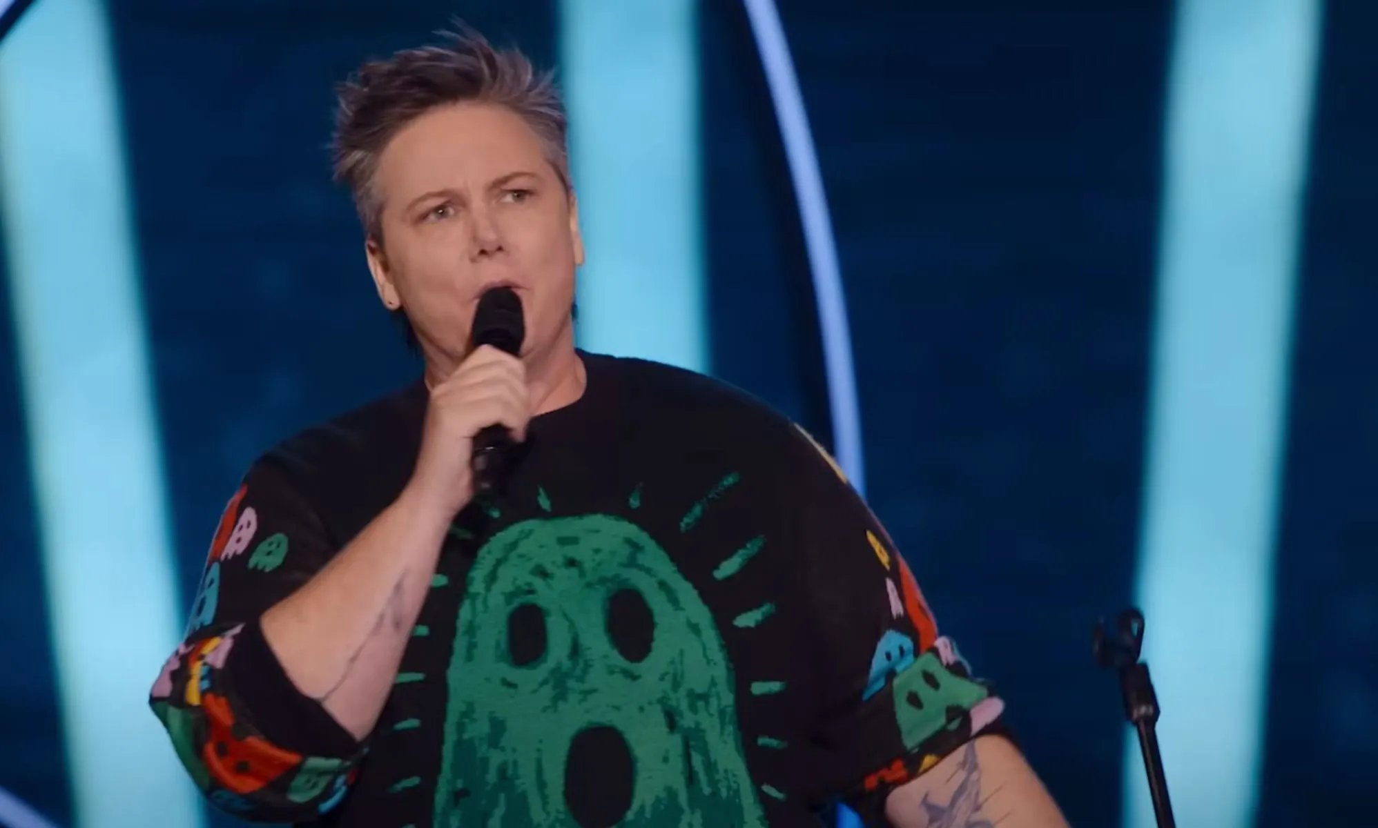 Australian comedian Hannah Gadsby wears a black shirt with a green ghost on it while speaking into a microphone during their new trans-led Netflix special Gender Agenda