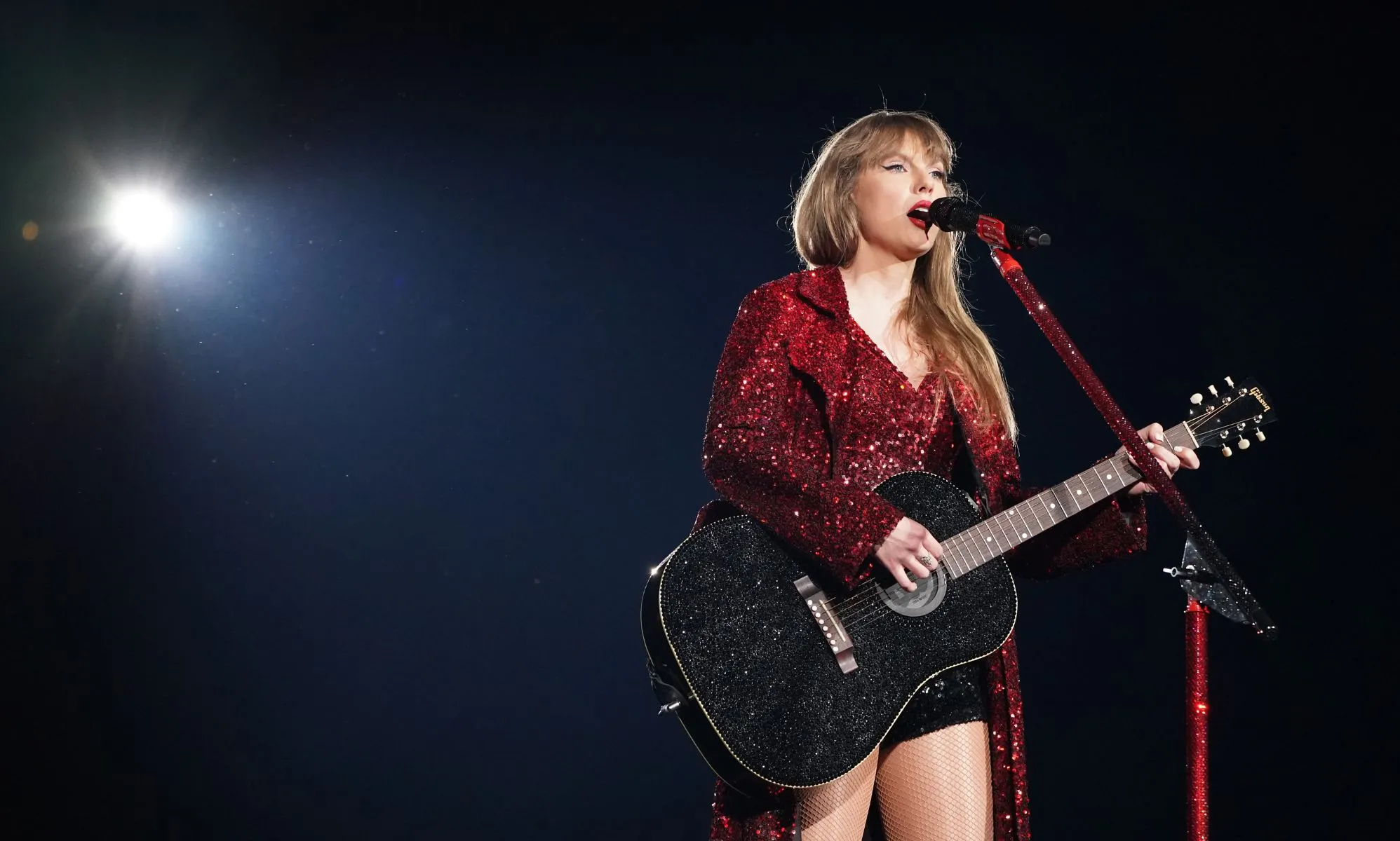 Taylor Swift wears a red sparkly outfit as she holds a guitar and sings into a microphone during a performance