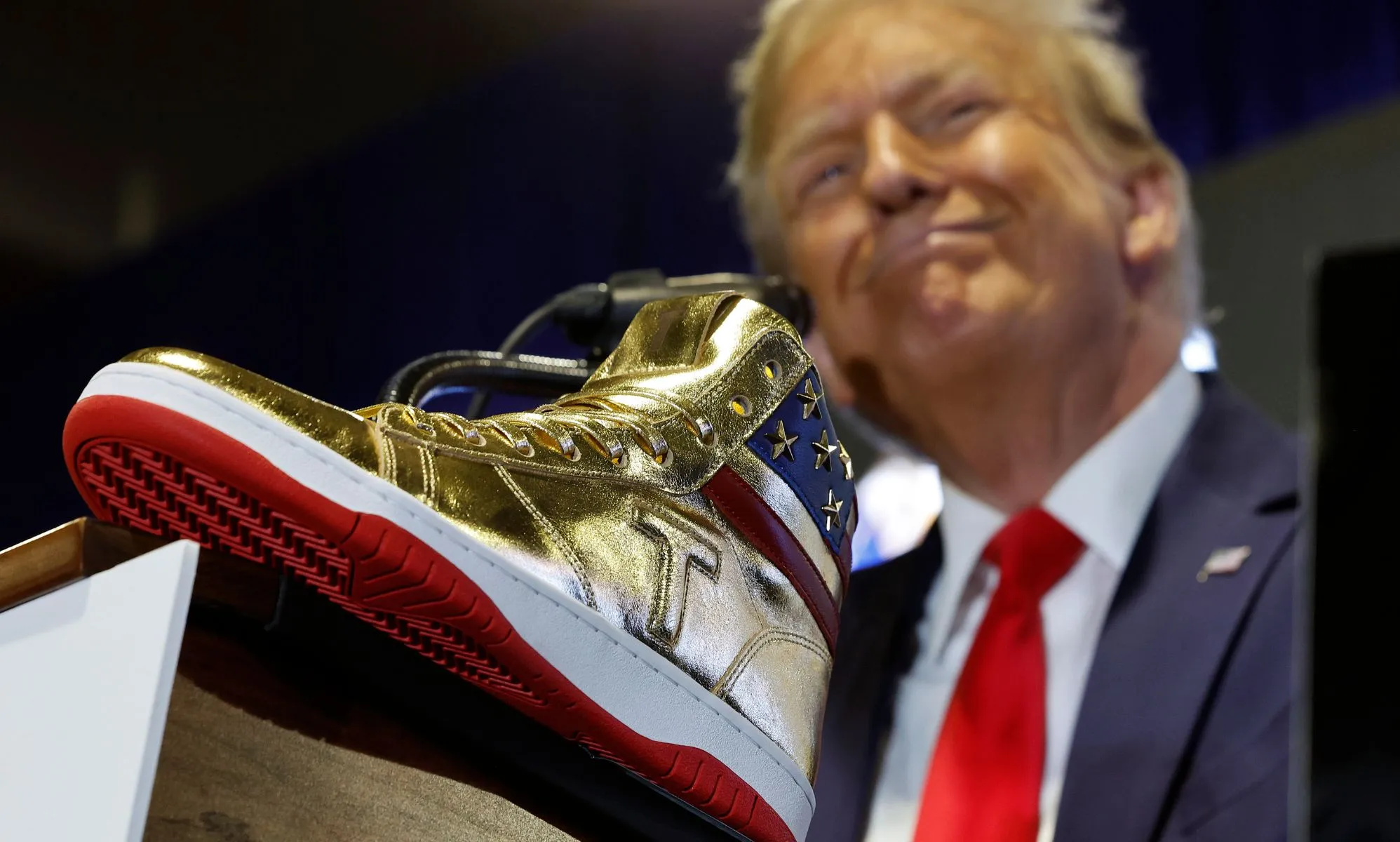 Donald Trump wears a suit and tie while smiling and standing in front of a gold-coloured high top sneaker with a T emblazoned on it and a US flag print