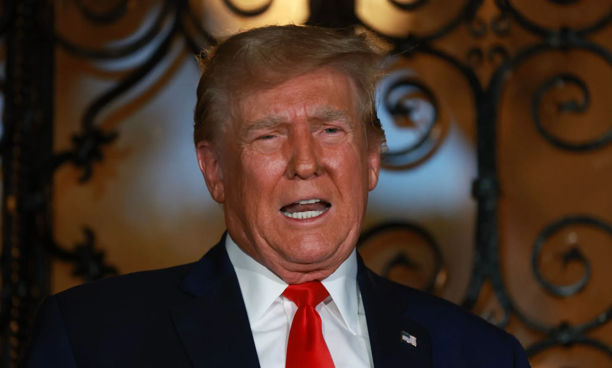 Former president Donald Trump wears a suit and tie and he appears before a door