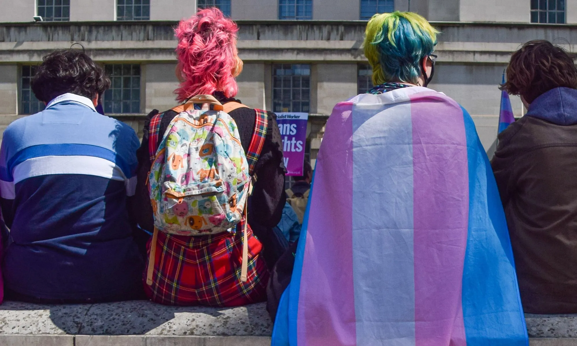 A group of people sit with their backs to the camera, one wearing a trans flag as a cape.
