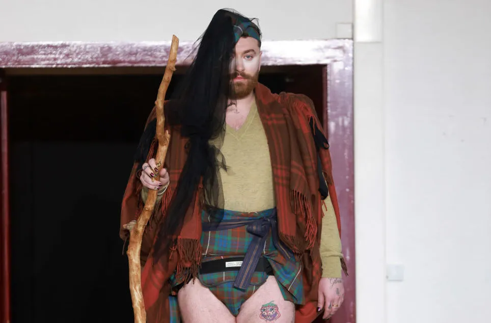 Non-binary singer Sam smith pictured at Paris Fashion Week wearing a tartan shall, a mini kilt, with long hair extensions and carrying a wooden staff