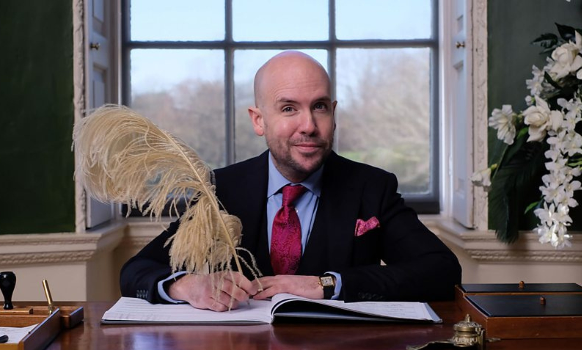Tom Allen sat at a wooden desk, a window behind him, signing wedding documents with a very large quill.