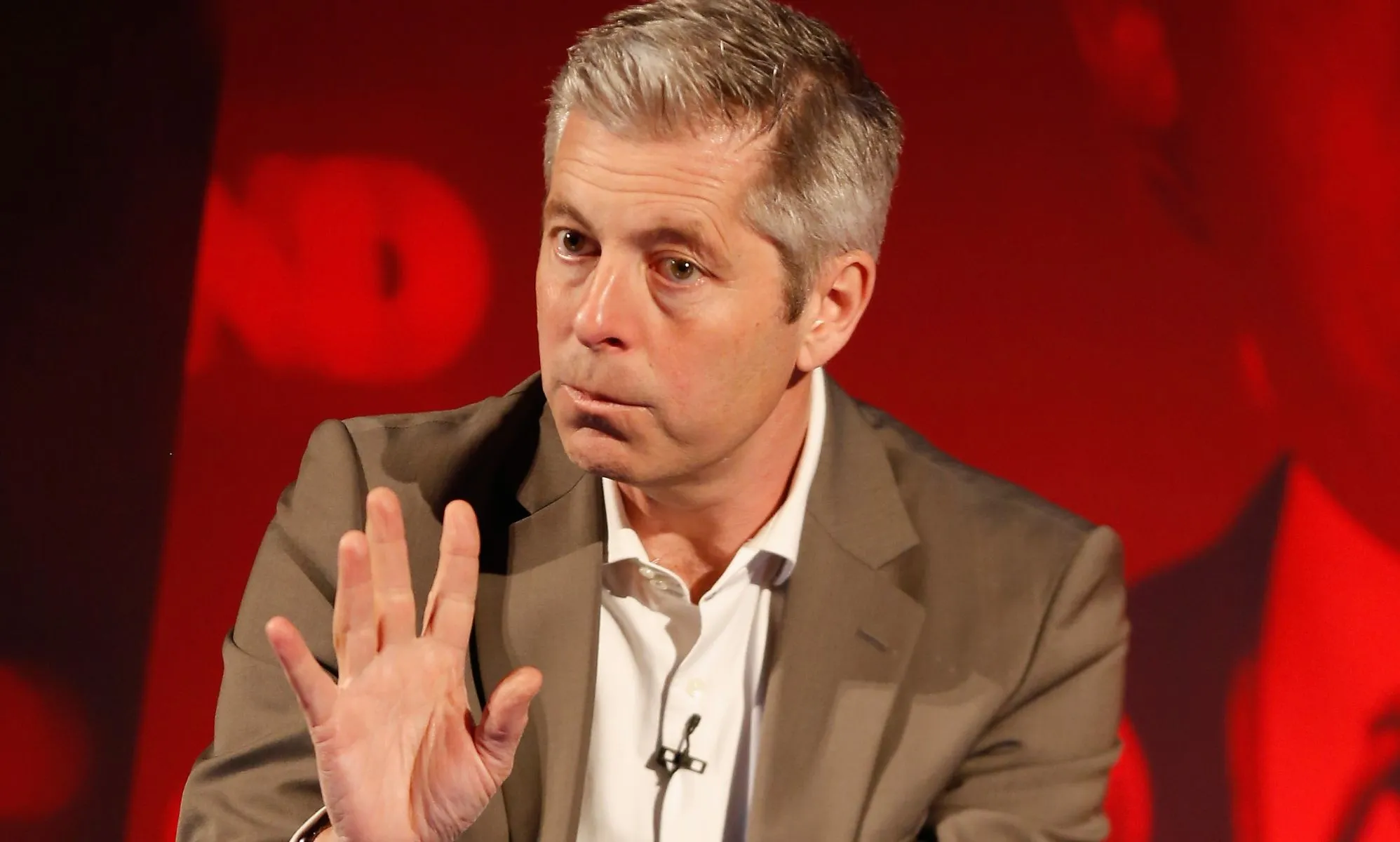 Justin Webb waving his hand during a speech on stage at a press event.