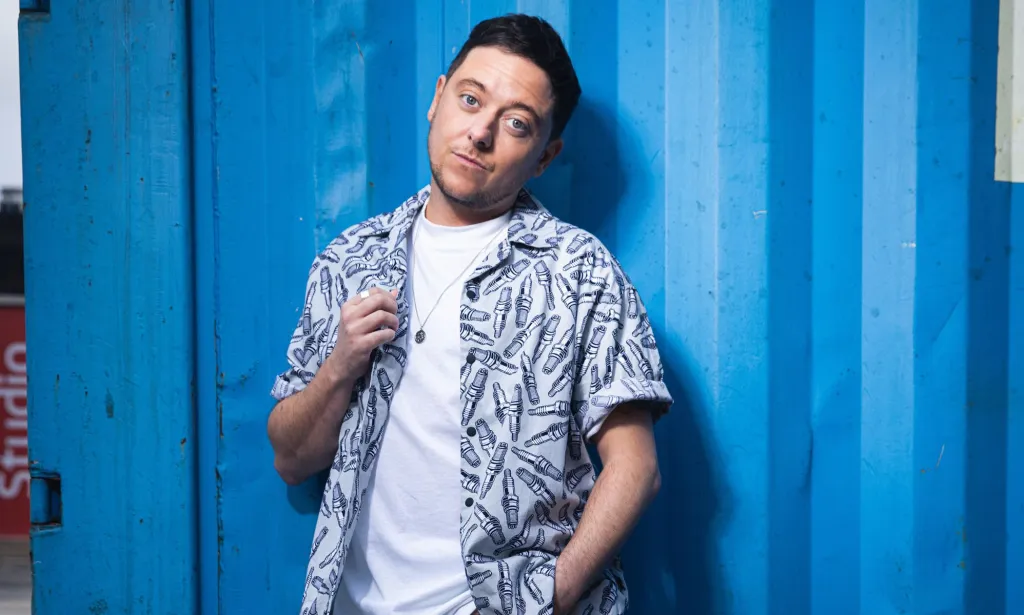 La estrella de Emmerdale y el actor trans Ash Palmisciano viste una camisa blanca y un botón estampado azul y gris mientras mira fijamente a la cámara.