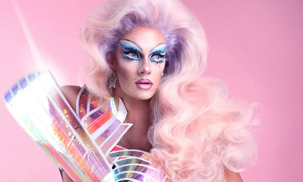 Una persona drag que lleva una peluca rosa posa frente a un fondo rosa.