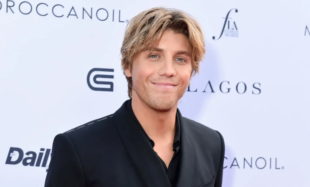 Lukas Gage in a black suit and shirt with parted blonde hair, smiling at the camera.