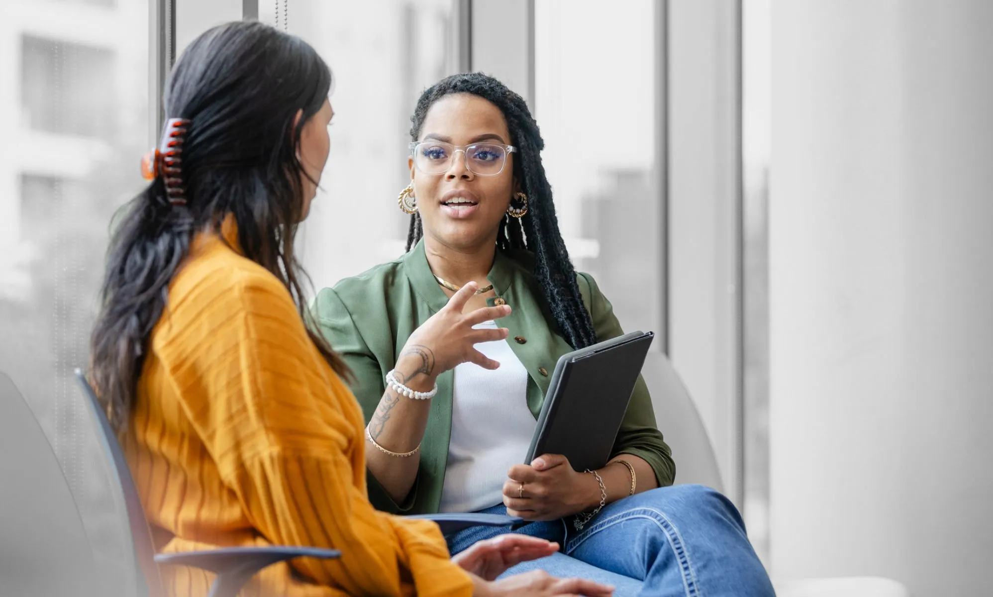 This is an image of two women talking. In the background there is a Black woman, wearing a green blazer, white tshirt and jeans. She is talking to someone wearing a yellow dress with long dark hair.