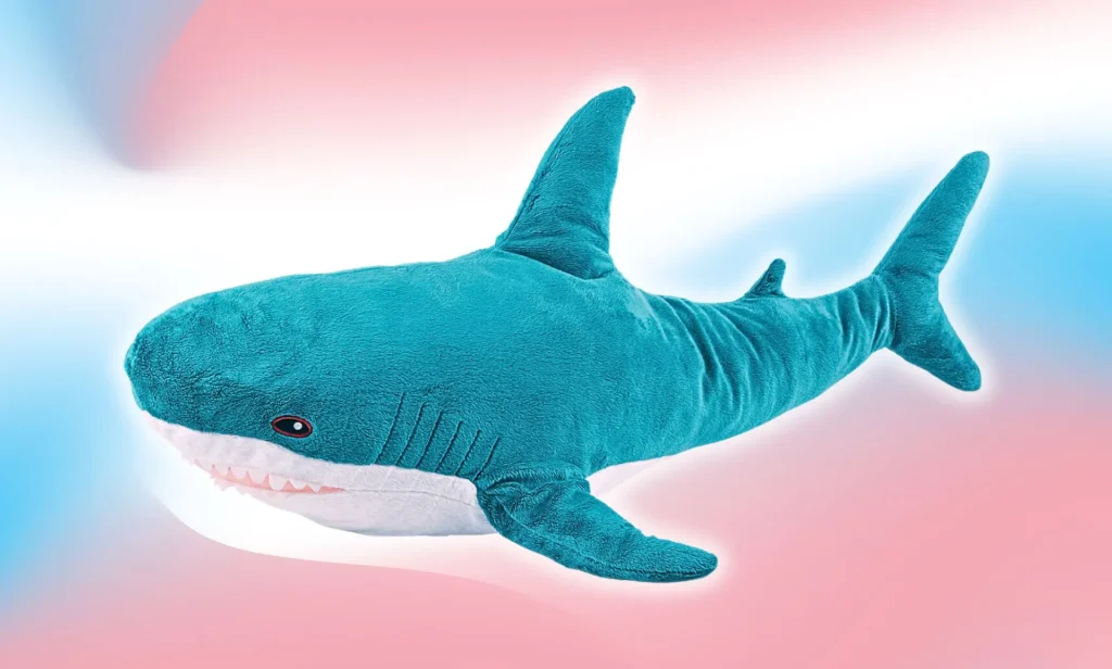 A Blahaj shark plushie photoshopped infront of a trans gradient.