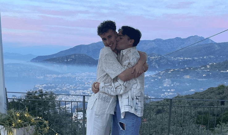 La foto muestra a Bradley Riches abrazando a su prometido con un patrón en forma de corazón hecho de rosas, contra un fondo tropical en un balcón.