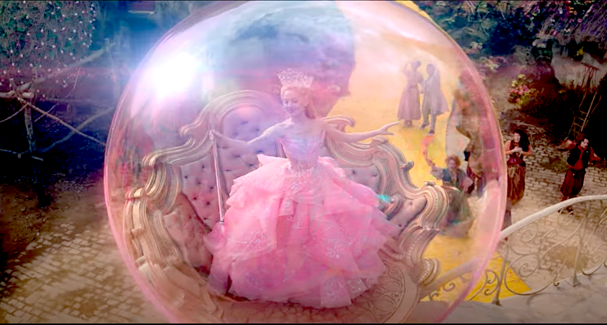 Photo shows Ariana Grande as Glinda The Good Witch floating in a pink bubble wearing a pink dress and holding a wand