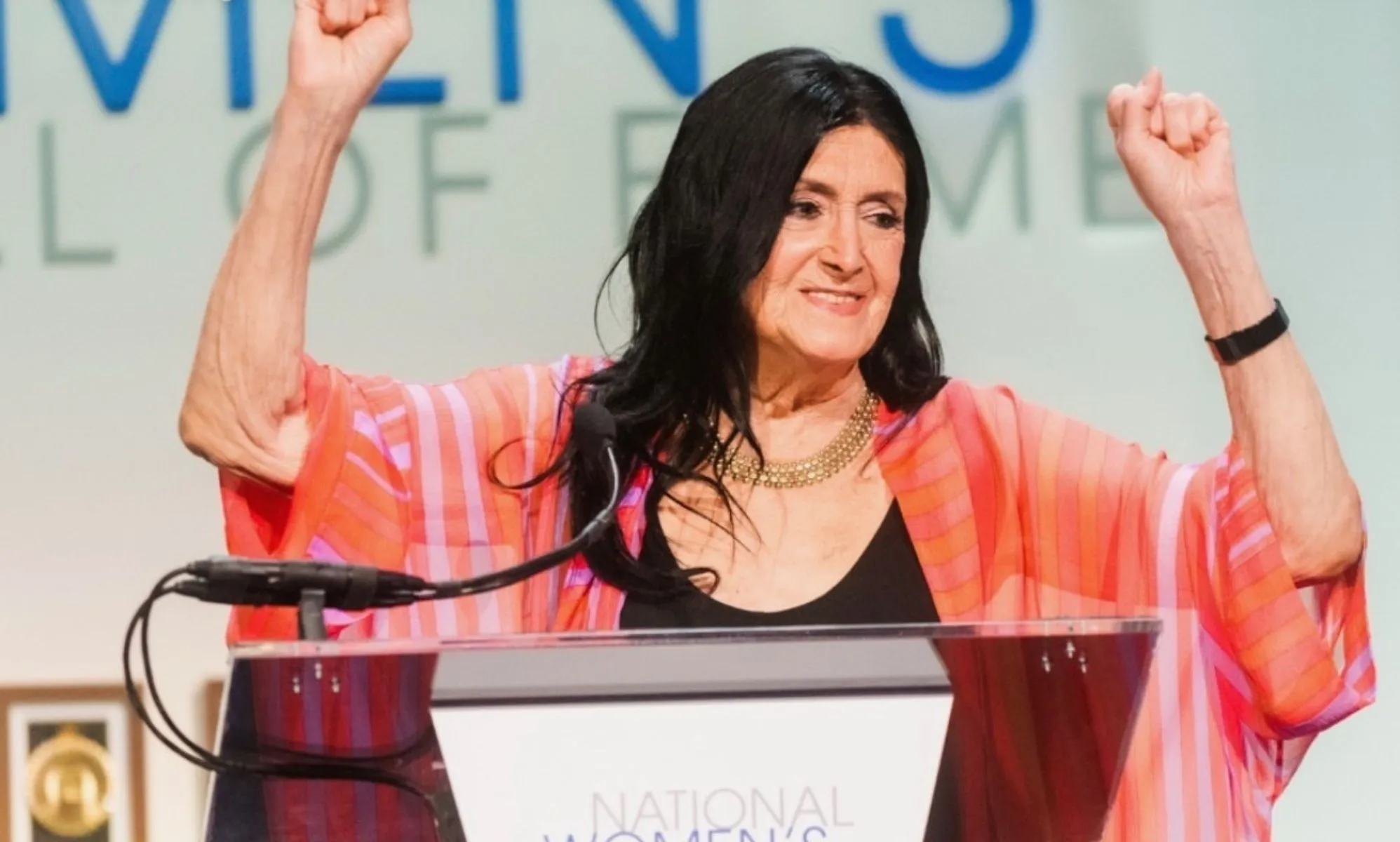 Trans academic and activist Sandy Stone wears a dark shirt and pink top as she holds up her hands while being inducted into the US National Women