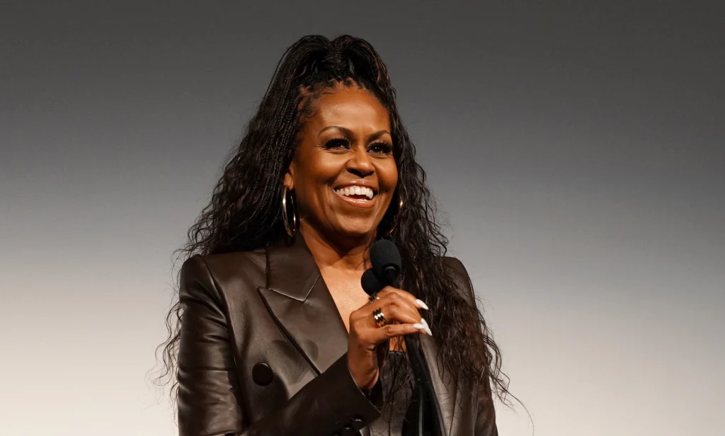 Michelle Obama with a mic smiling in a leather jacket with her hair up.