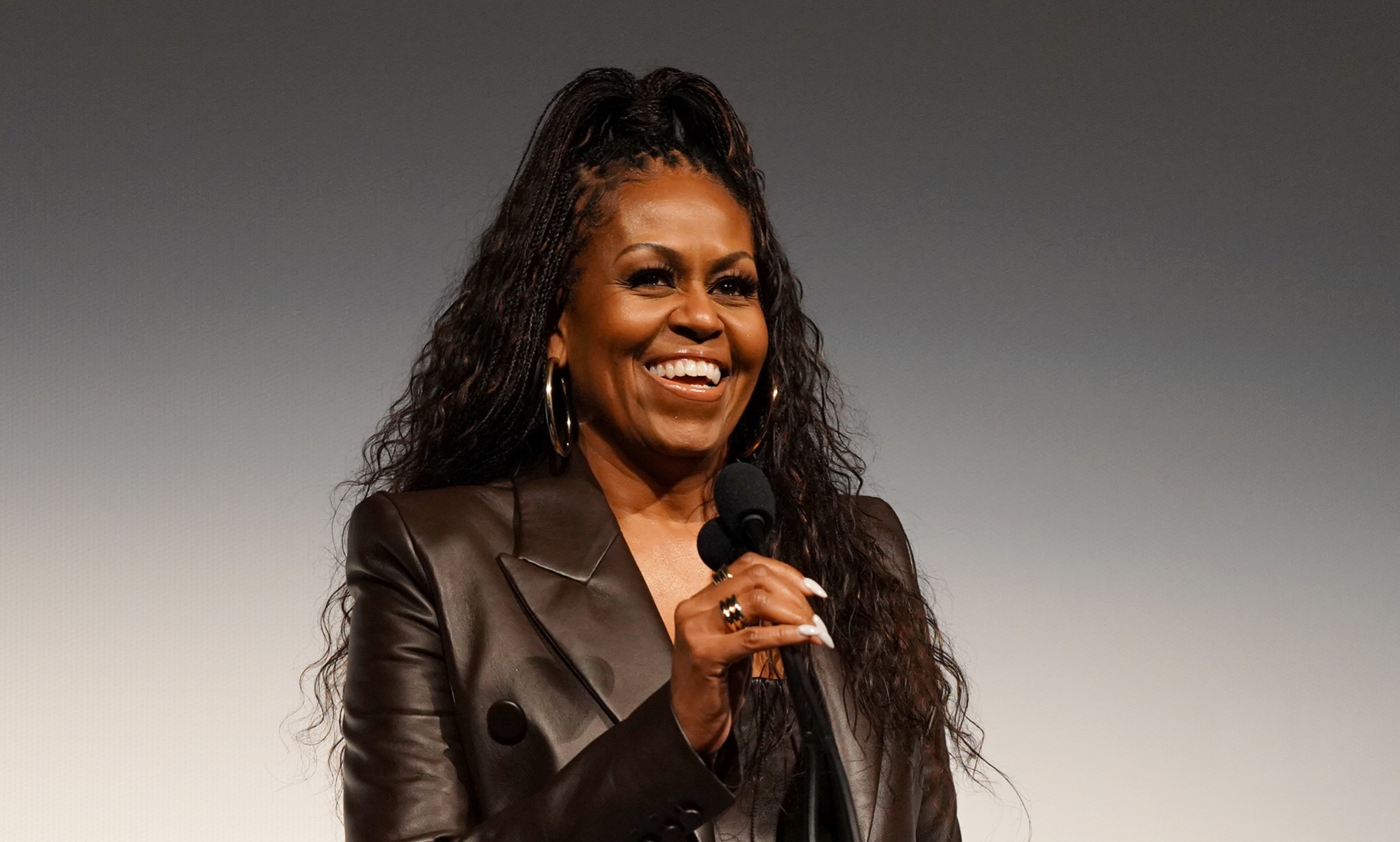 Michelle Obama with a mic smiling in a leather jacket with her hair up.