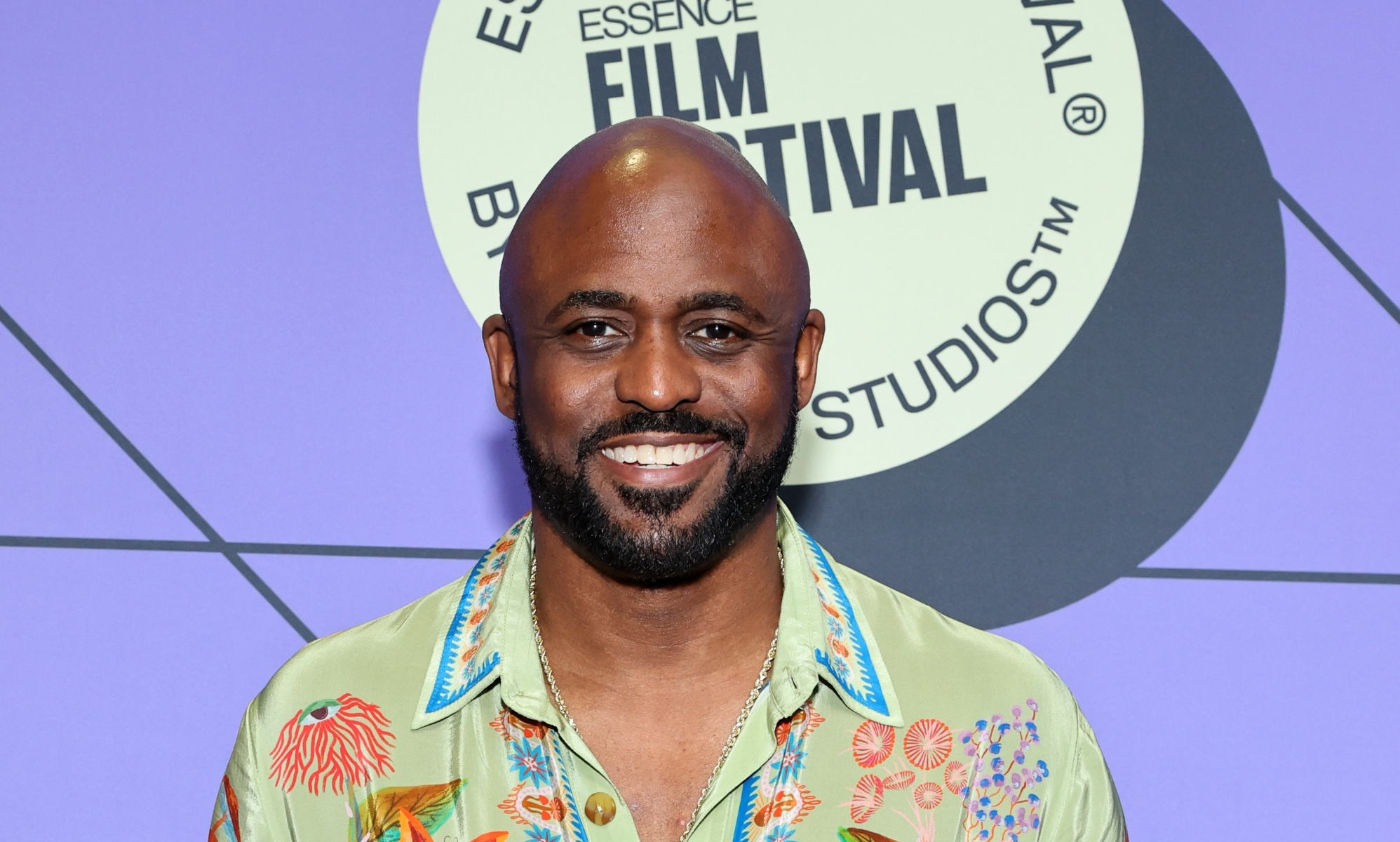 Wayne Brady smiling on a red carpet against a purple background and a floral design green shirt.