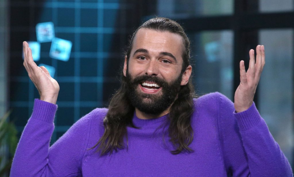 Jonathan Van Ness smiles and shrugs with their hands in the air.