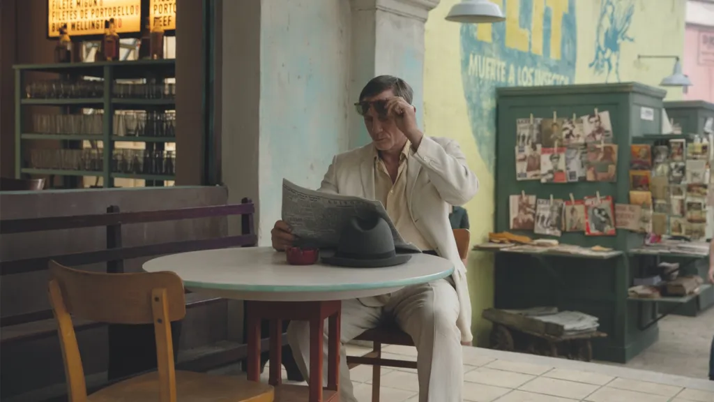 Aún más extraño, un hombre está sentado con un traje color crema, quitándose las gafas de sol mientras lee el periódico.