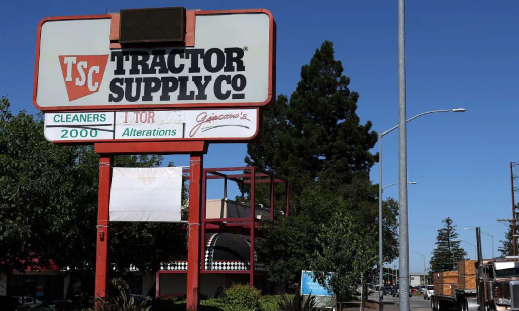 Esta es una imagen de un letrero de la tienda Tractor Supply Co. Está en un entorno rural y hay grandes pinos en el fondo.
