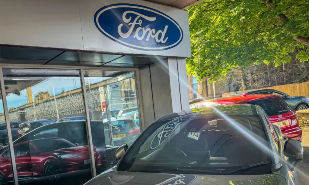 Esta es una imagen de un concesionario de automóviles Ford. El icónico logotipo de FORD en azul con letras plateadas se encuentra sobre un edificio de vidrio. También hay algunos automóviles en la foto.