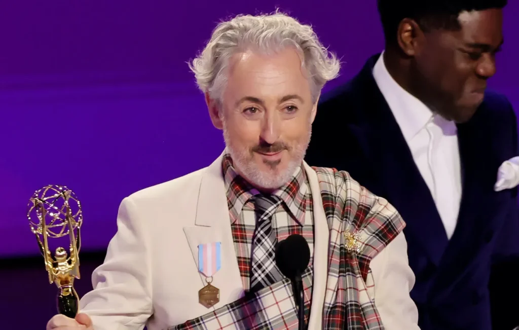 Alan Cumming on stage at the Emmys, holding his award, close cropped to show the trans pride flag medal