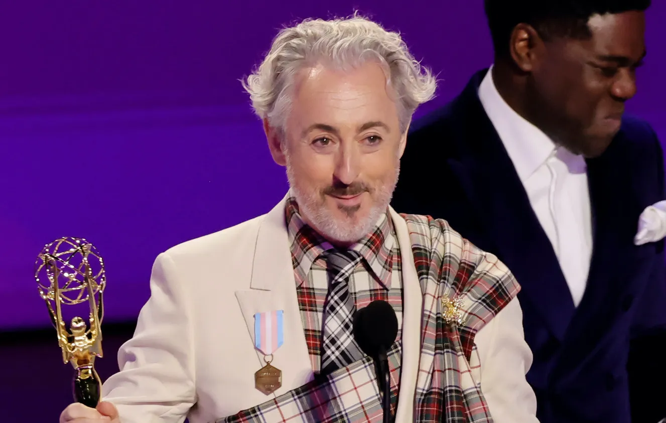 Alan Cumming on stage at the Emmys, holding his award, close cropped to show the trans pride flag medal