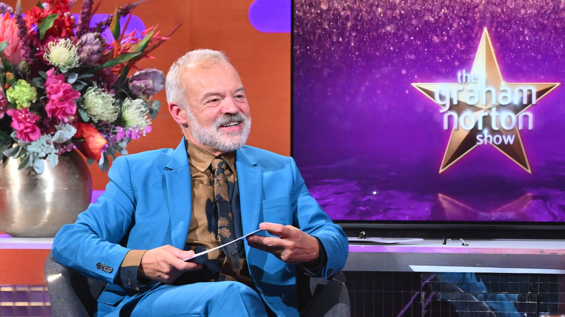 The Graham Norton Show, Graham Norton, in a blue suit, sitting next to a screen with the show name.