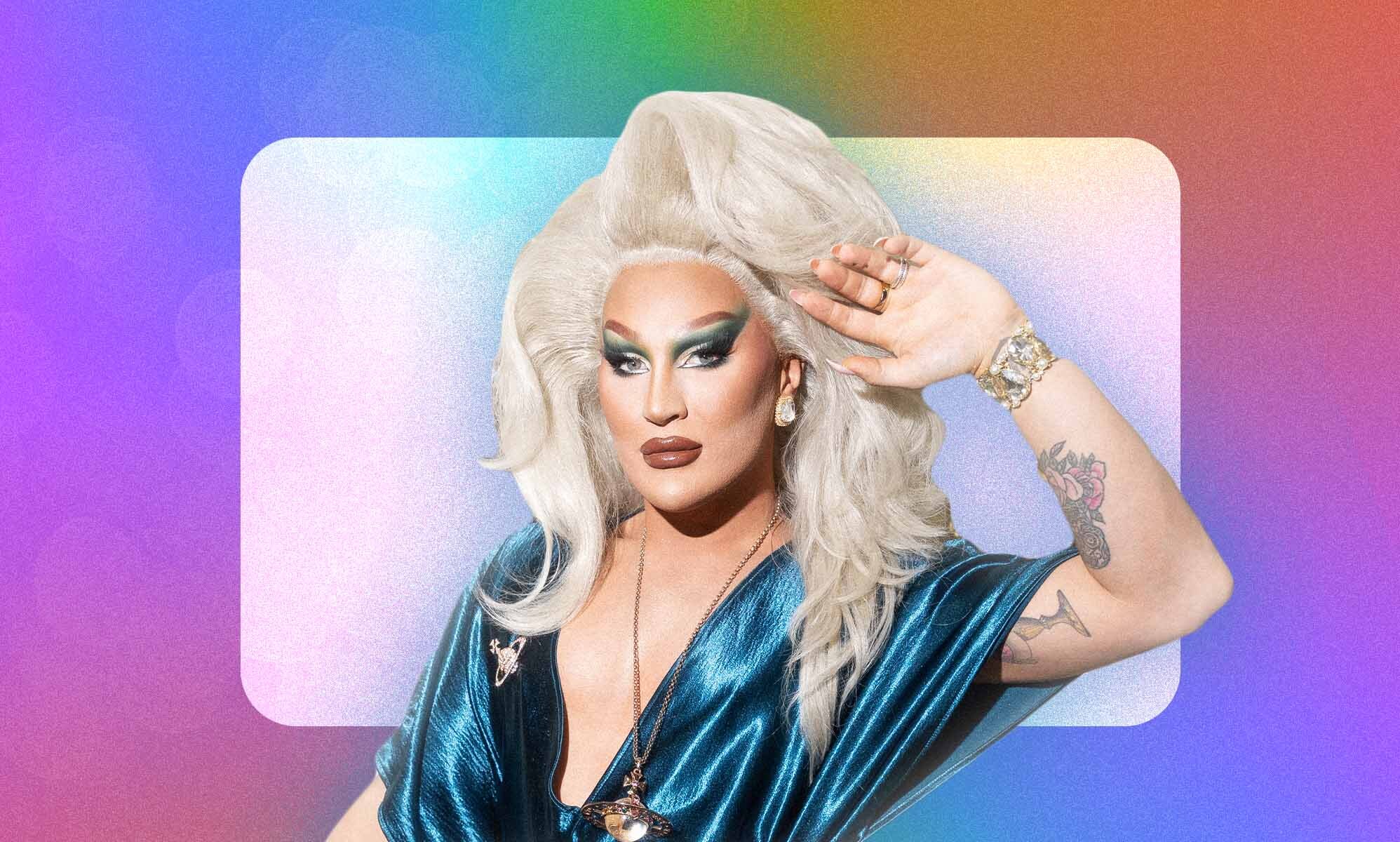 The Vivienne in a blue dress holding her arm against her blonde wig against a colourful rainbow background.