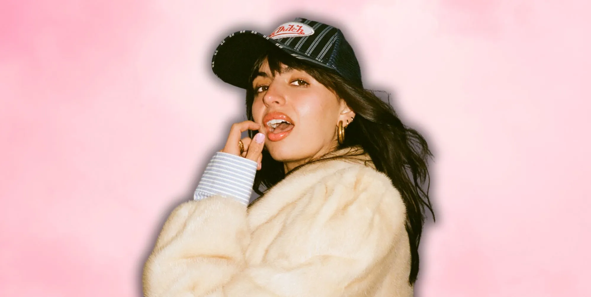Rebecca Black in a white faux fur jacket and baseball cap, posing with her mouth open and finger and thumb near her face.