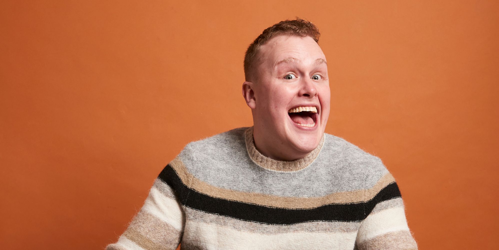 Comedian Josh Jones beams with his mouth open. He is in a grey black and beige striped jumper and sitting against an orange background.