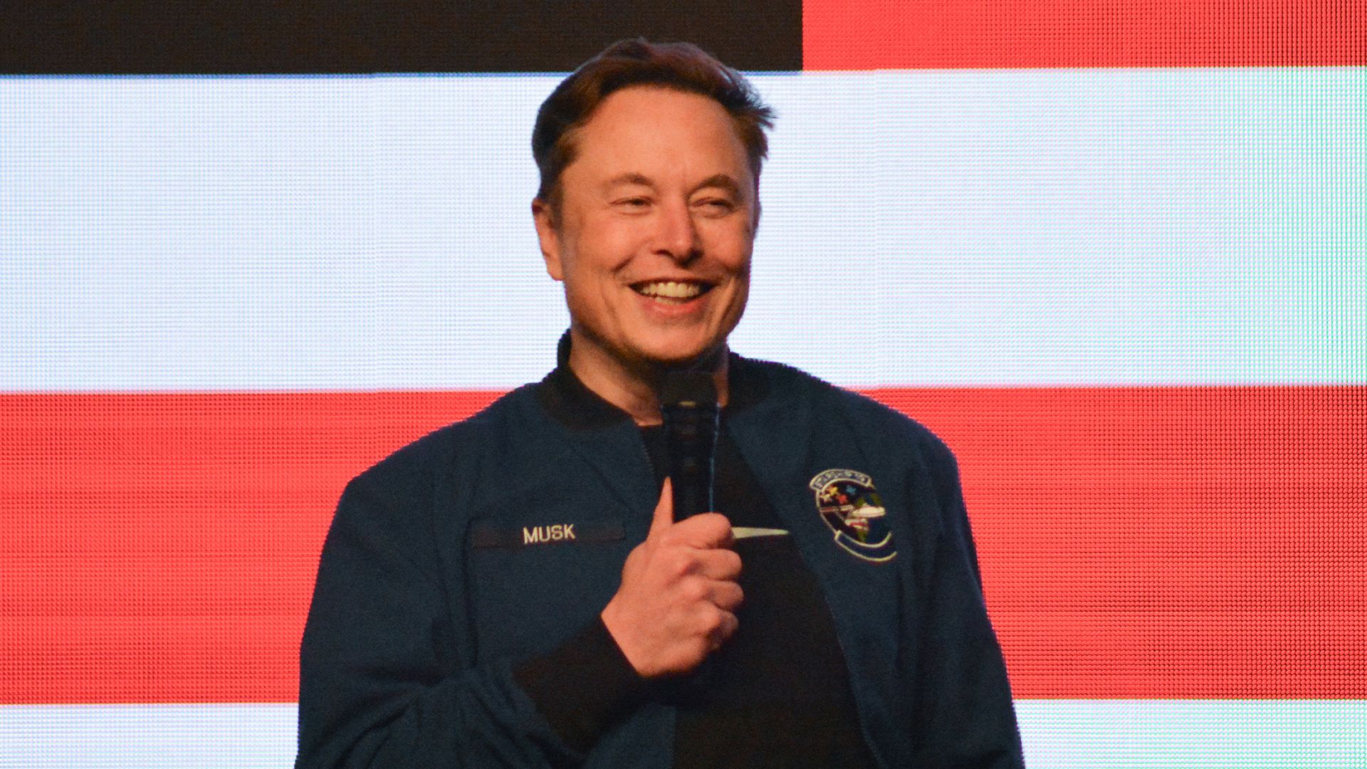 Elon Musk stands with a microphone in front of an American flag background