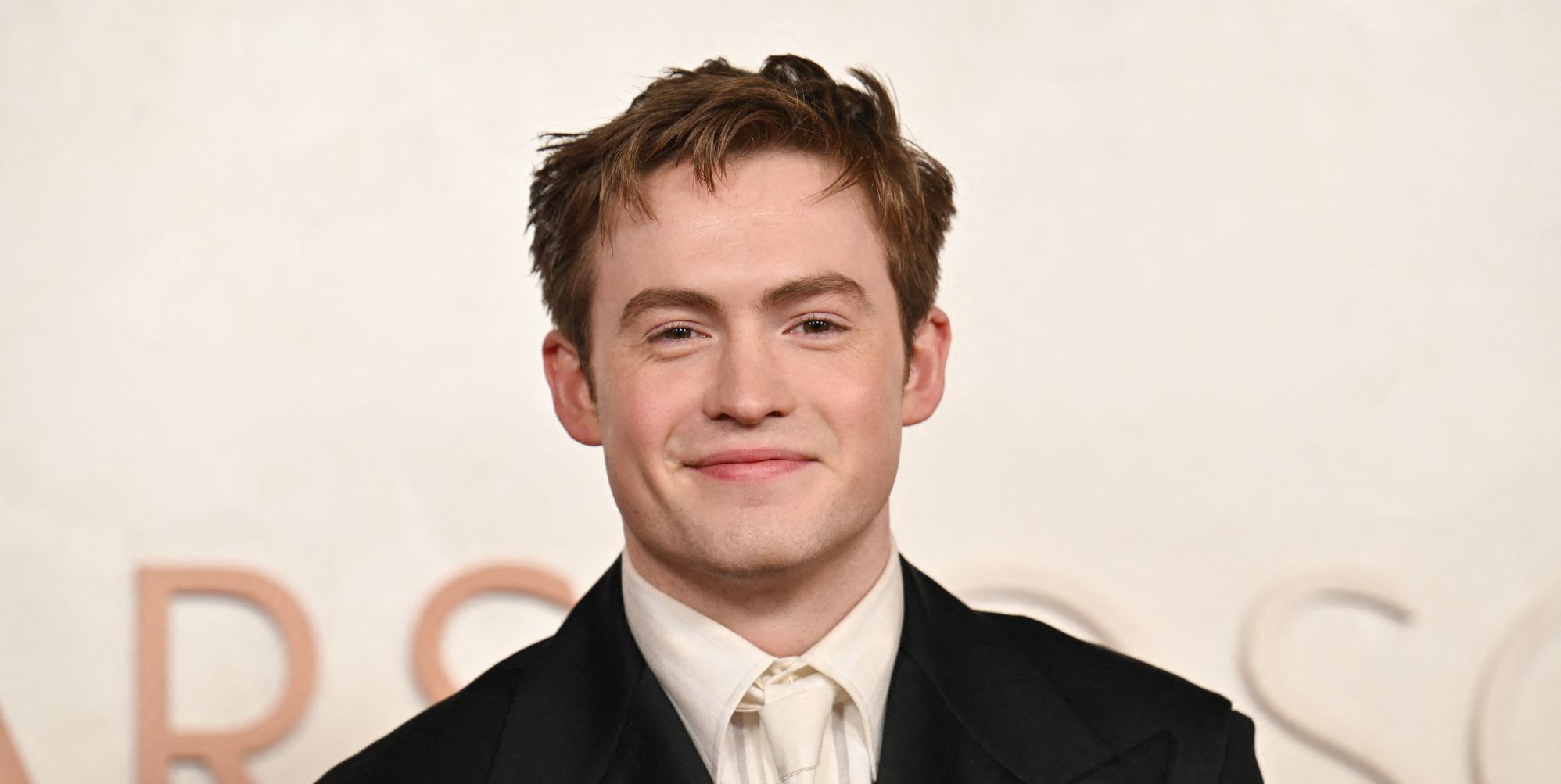 Kit Connor smiles at a red carpet event while wearing a cream t-shirt, black blazer, against a cream background.