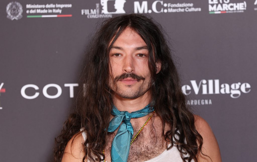 Ezra Miller attends Filming Italy 2025 They have long wavy brown hair over their shoulders. A silk blue necktie, gold beaded necklace and white vest.