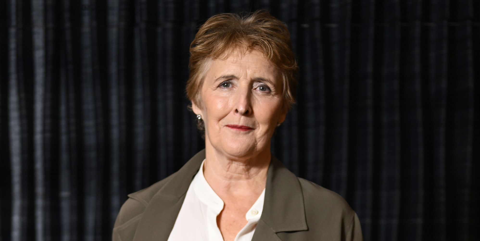 Fiona Shaw in a grey jacket, white shirt, against a black background.
