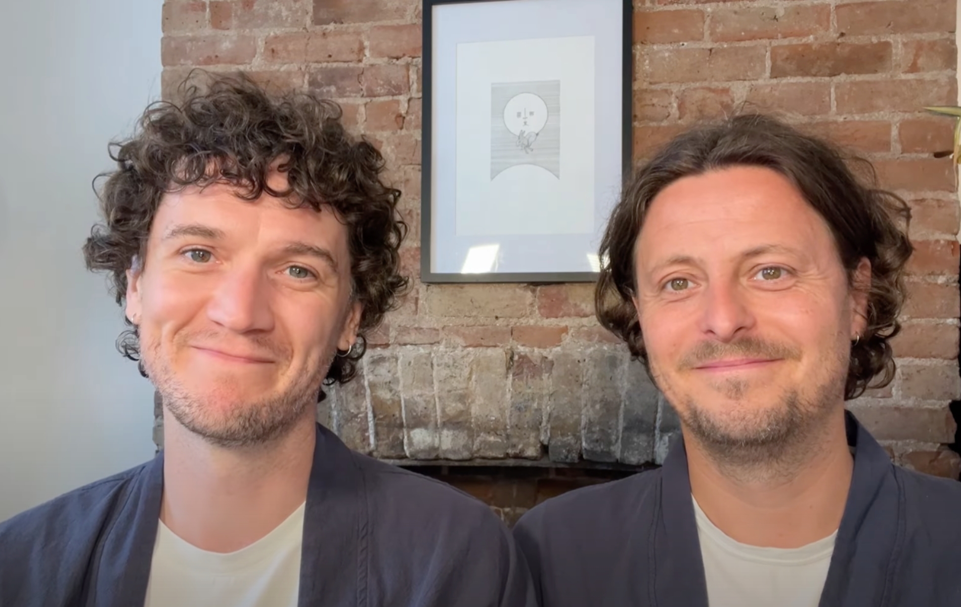 Fiongal and Jamie Greenlaw-Meek both wearing white t-shirts and grey over shirts sat against a brick wall with a moon painting.
