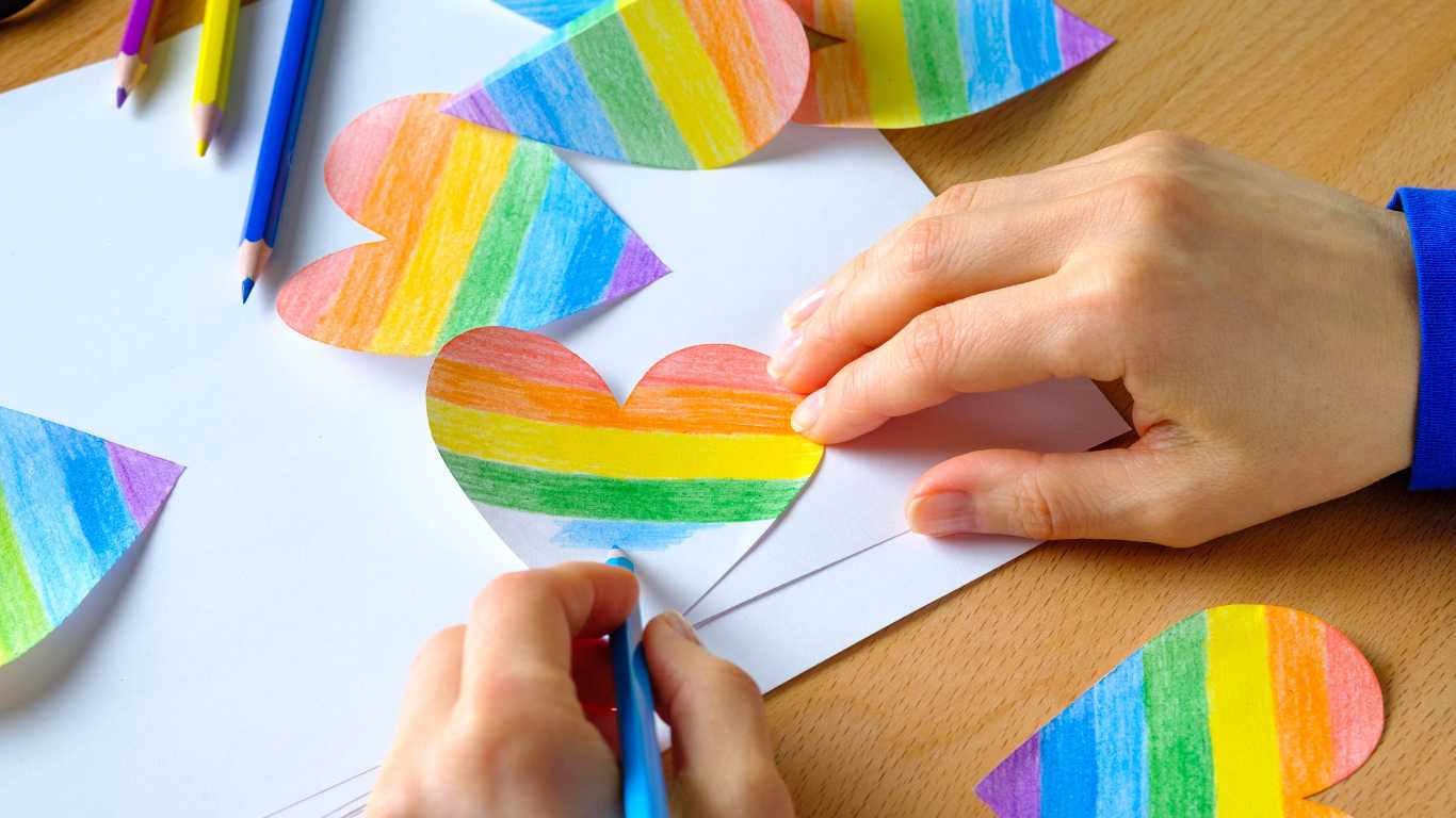 A girl colours a Heart cut out of paper in the colours of the LGBTQ Pride flag.