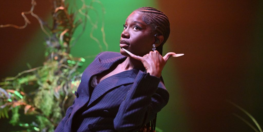 Doechii leans back while wearing a suit and makes a phone gesture with her hand.