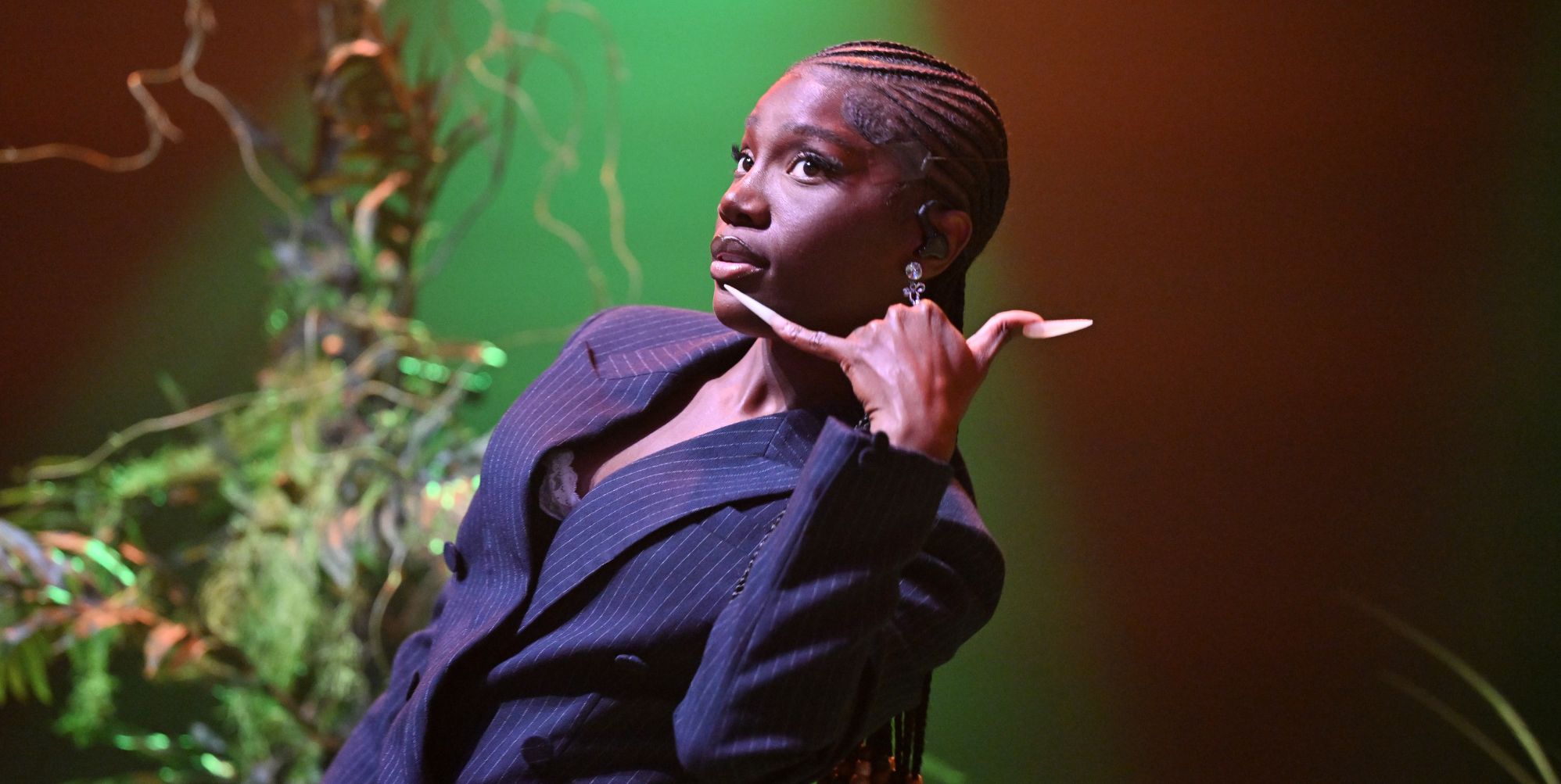 Doechii leans back while wearing a suit and makes a phone gesture with her hand.