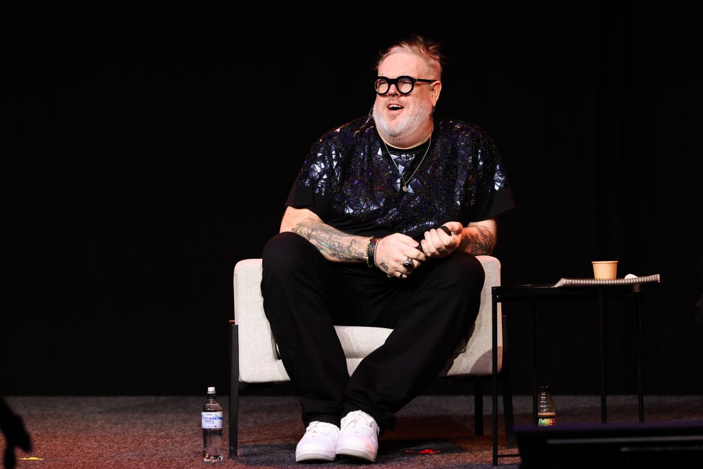 Kristian Nairn se sentó en una silla blanca en un panel. Está sosteniendo un micrófono y vistiendo una camisa reflectante