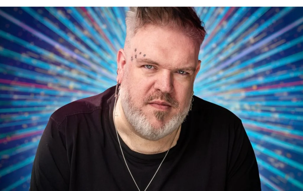 Kristian Nairn wearing a black t-shirt looking into camera with classic Strictly blue sparkles behind him.