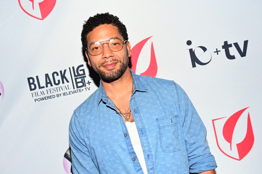 Jussie Smollett con una chaqueta de mezclilla sobre un chaleco blanco. También lleva gafas plateadas teñidas.