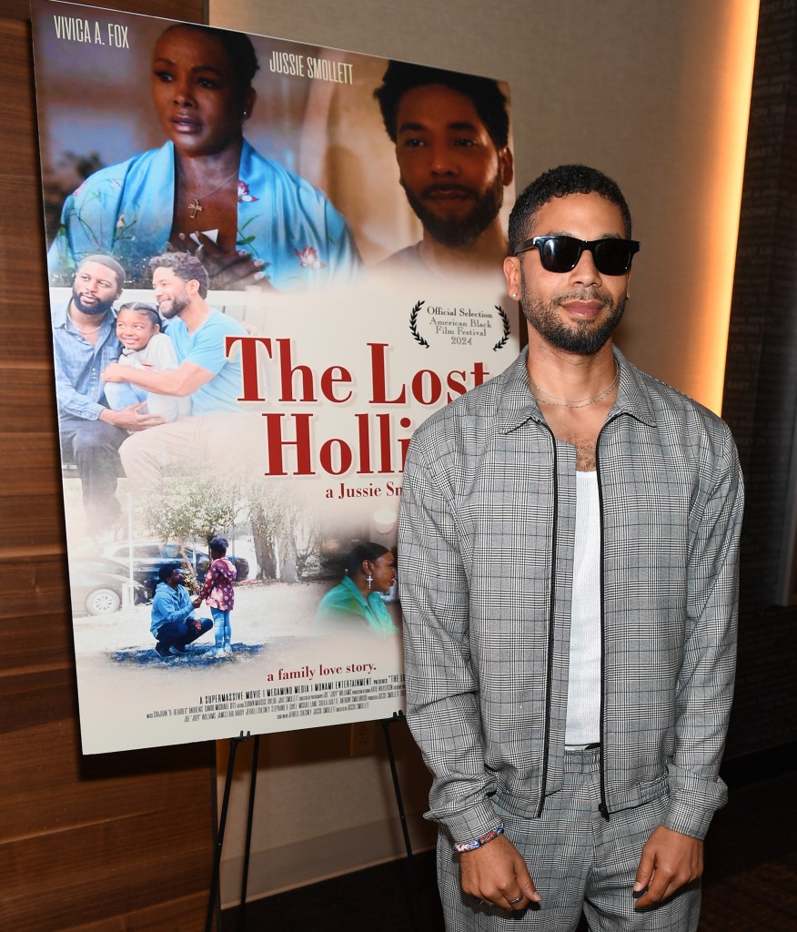 Jussie Smollett asiste al estreno de Atlanta de "El Holliday perdido. Está parado frente al póster de la película I na gris y gafas de sol negras.