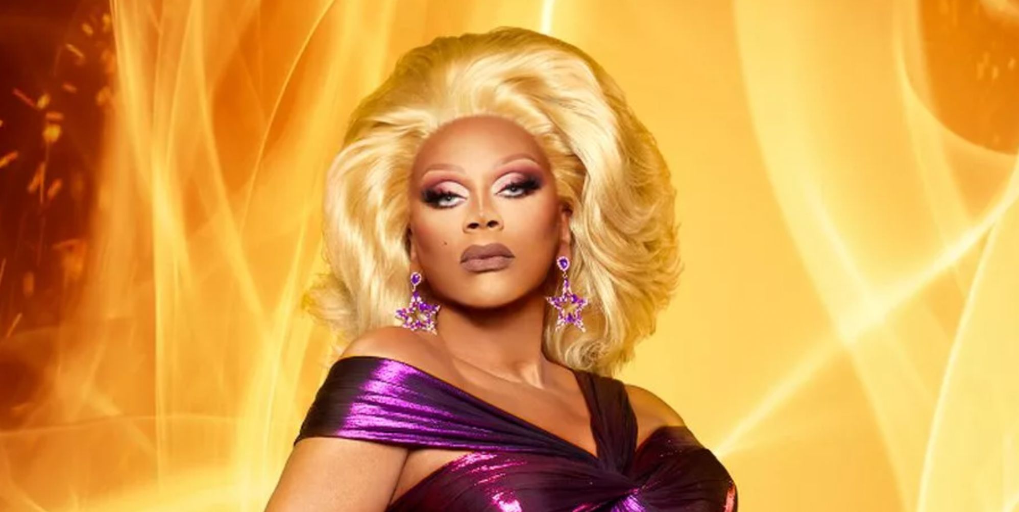RuPaul poses in a purple dress against a backdrop of flames.