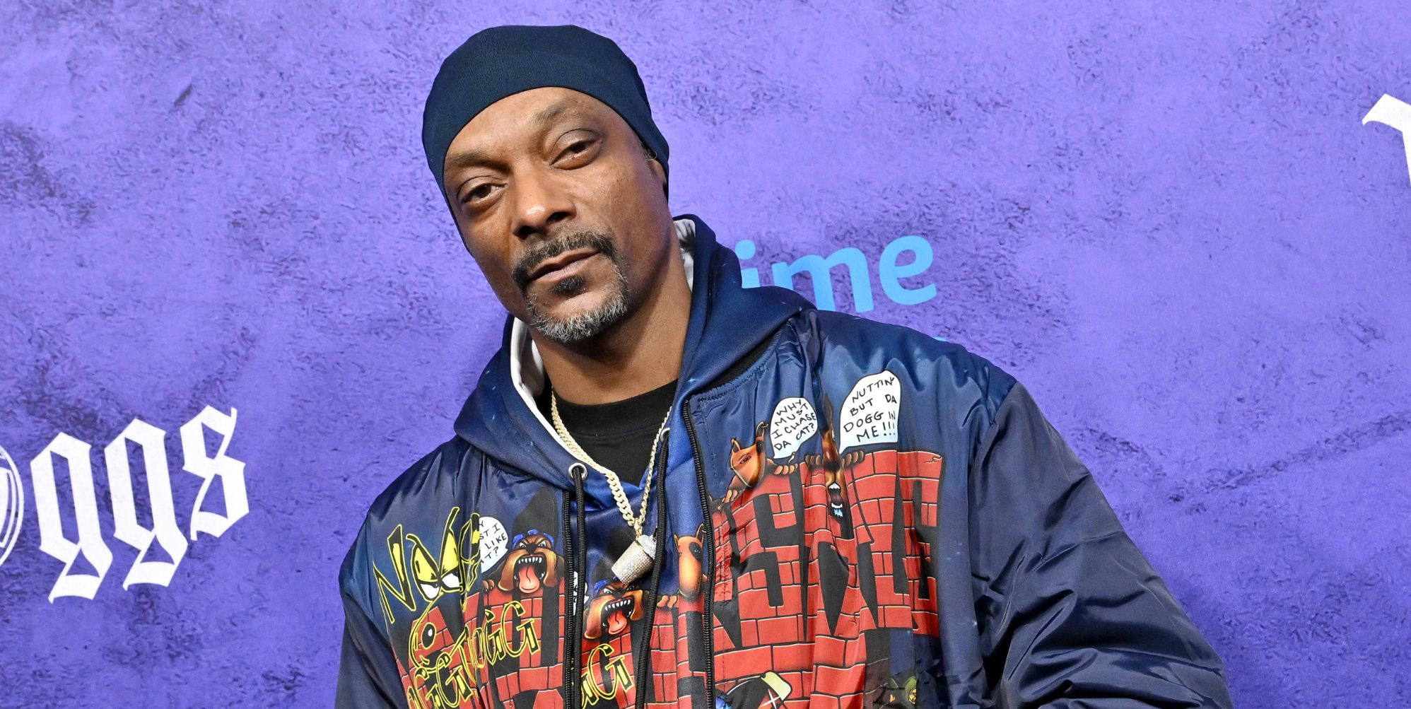 Snoop Dog in a blue yello and red denim jacket at a premiere with a purple background behind him.
