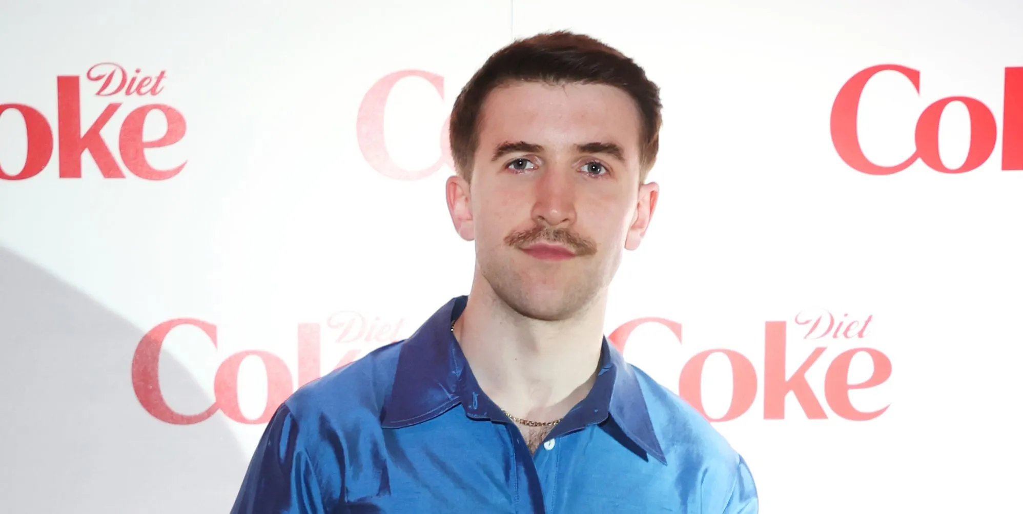 Callum Scott Howells poses in a blue shirt against a white background with Coke written on it.