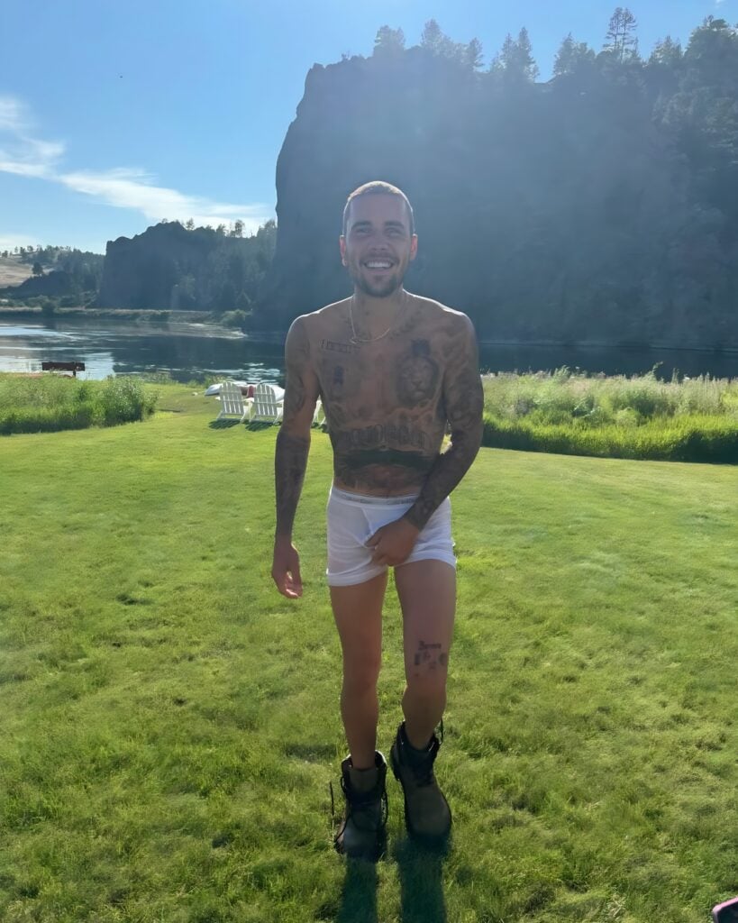 Justin Bieber Mid-stride en un césped soleado, con calzoncillos blancos húmedos y botas, sonriendo con un lago y un acantilado en el fondo.