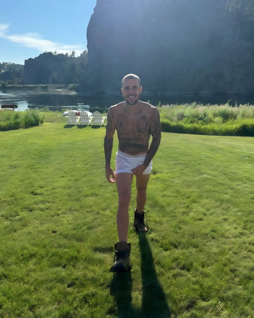 Justin Bieber camina hacia la cámara con calzoncillos blancos empapados y botas, sonriendo contra un pintoresco lago y un telón de fondo.