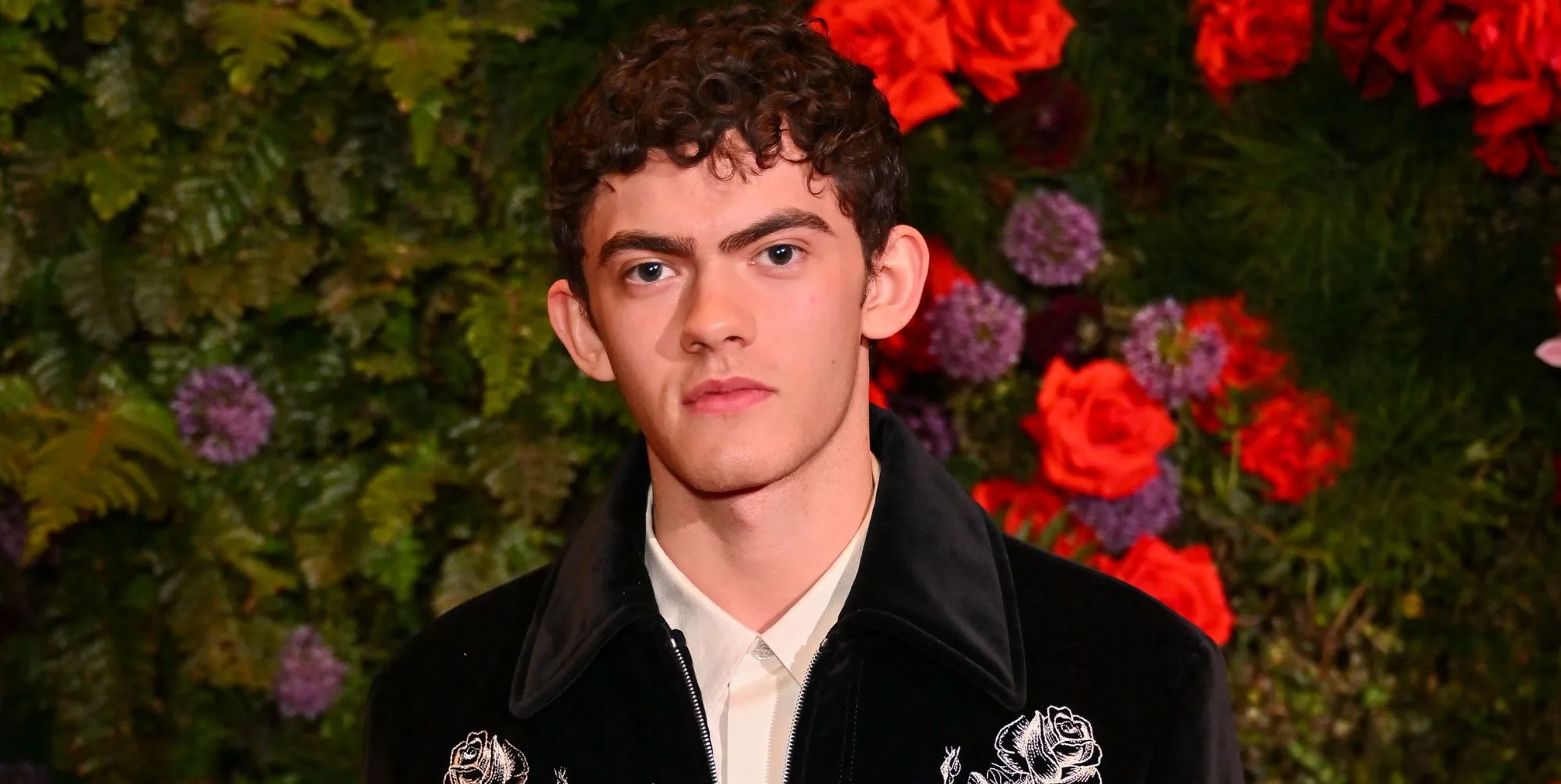 Joe Locke poses in a black jacket and white shirt against a flowery background.