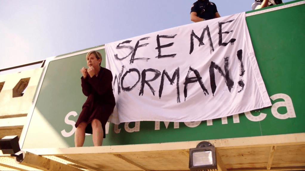 Una mujer se paró en un letrero sobre una autopista con una pancarta que decía 'See Me Norman'.