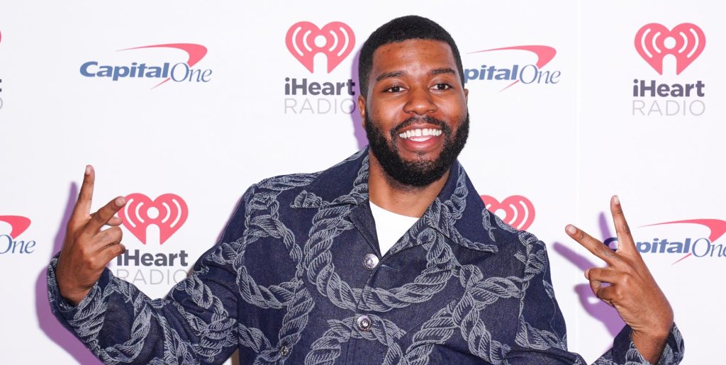Khalid gives a two fingered salute and smiles as he wears a rope patterned denim shirt while appearing at a red carpet event.